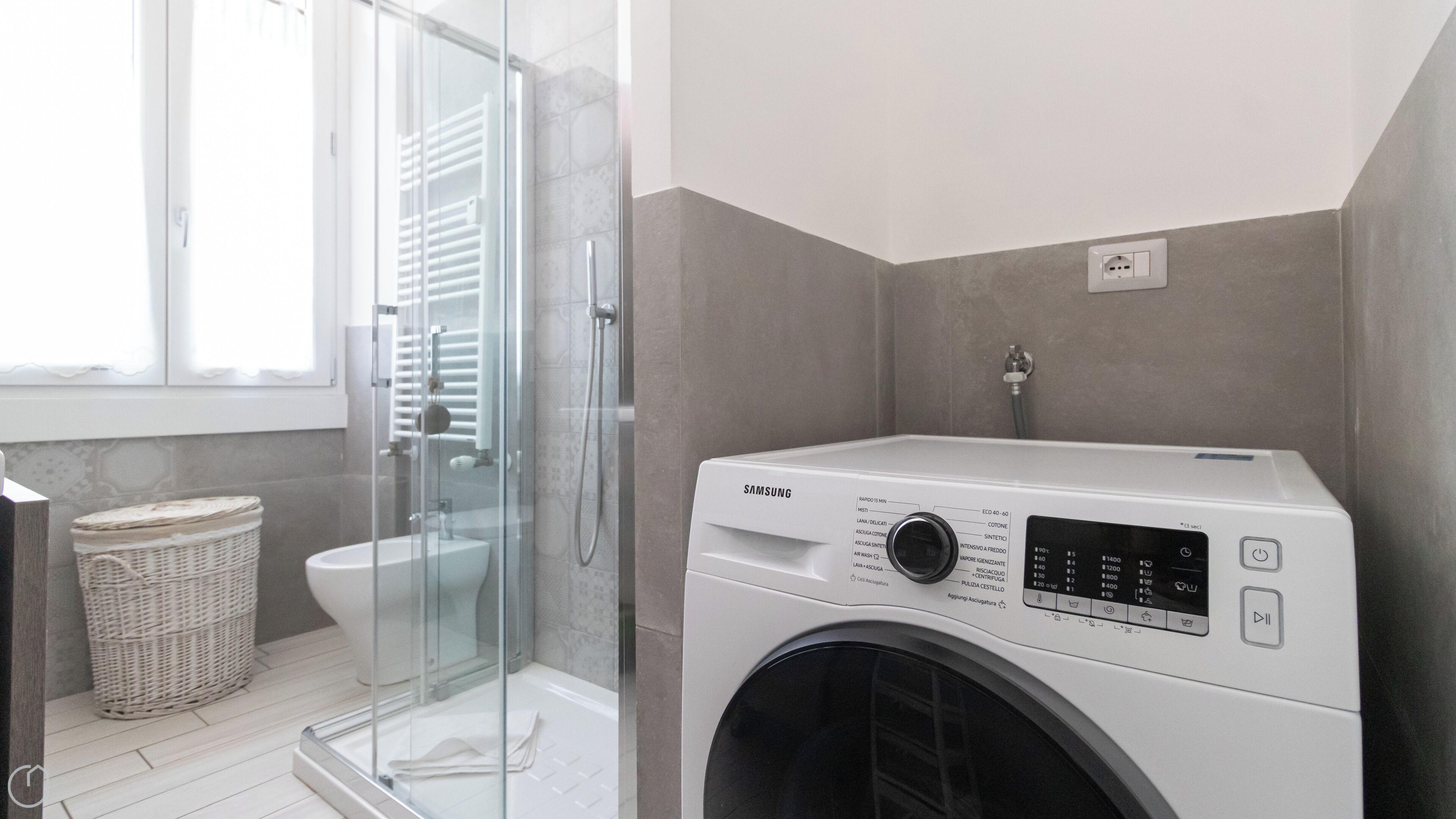 A modern bathroom with a glass shower enclosure, a white washing machine, and a wicker basket.