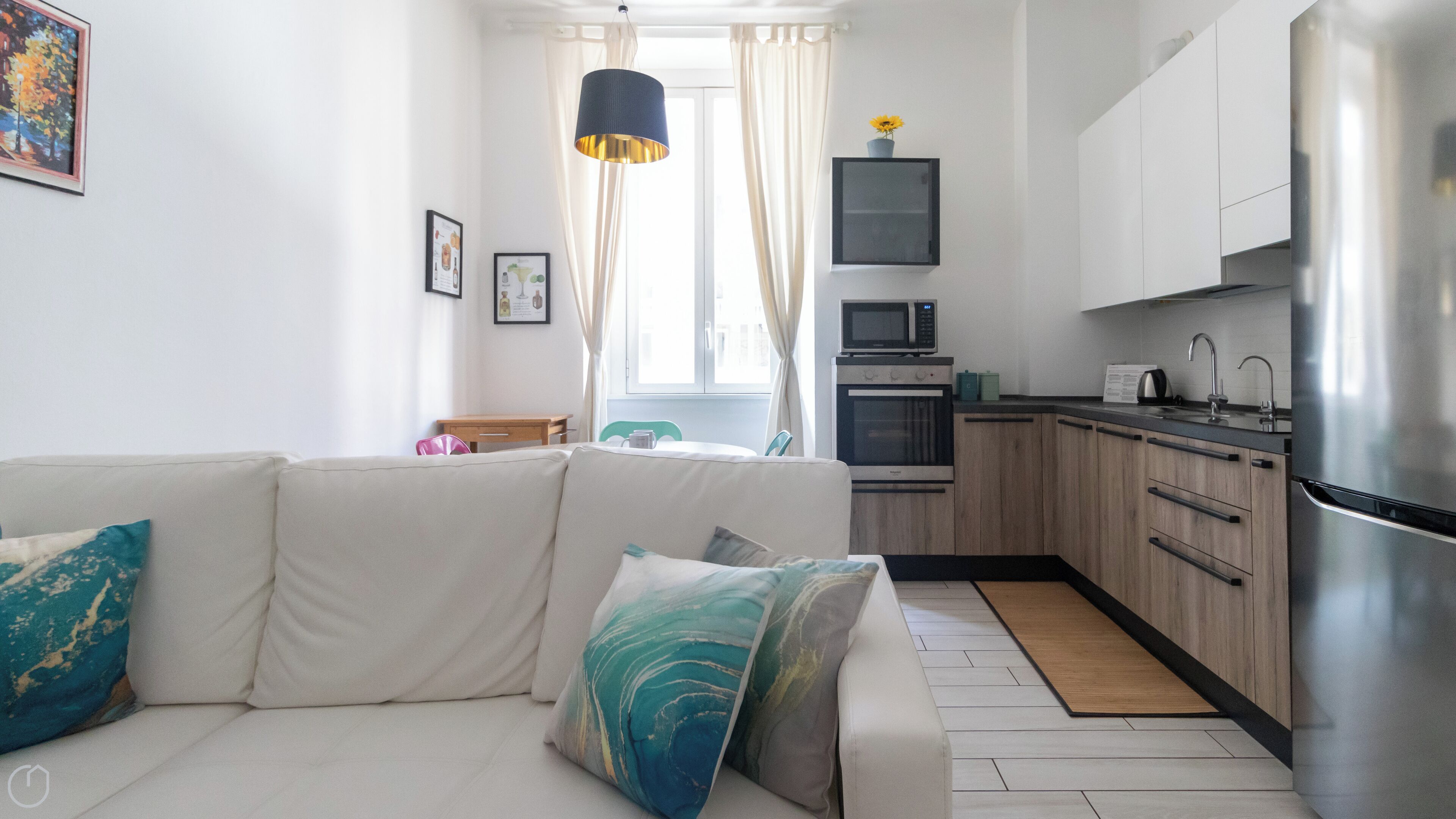 Un salon moderne avec un canapé blanc, une petite table et un coin cuisine doté de meubles en bois et d’appareils électroménagers en acier inoxydable.