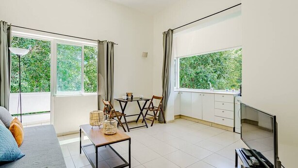 A modern living room with a sofa, a dining table with chairs, a TV, and large windows with curtains. - Central 1BR in Dizzengoff by Holiday-rentals (Tel Aviv-Yafo)