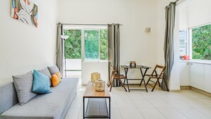 A modern living room with a grey sofa, a small table, and a dining area with chairs. - Central 1BR in Dizzengoff by Holiday-rentals (Tel Aviv-Yafo)