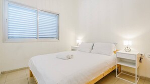 A bedroom with a wooden bed, white bedding, a nightstand with a lamp, and a window with blinds. - Central 1BR in Dizzengoff by Holiday-rentals (Tel Aviv-Yafo)