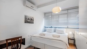 Une chambre à coucher comprenant un lit, un bureau, une chaise, une fenêtre avec des stores et un climatiseur fixé au mur.