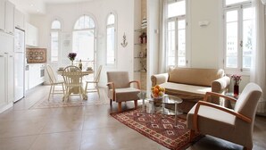 Een ruime woonkamer met een beige bank, twee fauteuils, een glazen salontafel en een eettafel met stoelen. Er zijn grote ramen die natuurlijk licht en uitzicht bieden.