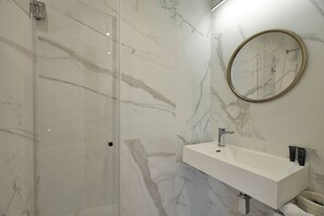 Un baño moderno con cabina de ducha de vidrio, pared con azulejos de mármol, lavamanos blanco con espejo y un perchero.