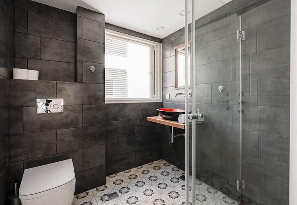 A modern bathroom with dark tiles, a toilet, a sink with a red basin, and a shower area. - Unique 2BR Frishman by Holiday-rentals SAFE ROOM (Tel Aviv-Yafo)