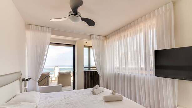 A hotel room with a bed, a flat-screen TV, a ceiling fan, and a view of the ocean through the window. - Wonderfull duplex 3BR Hakovshim SAFE ROOM ({{addressCityy}})