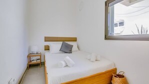 Ein ordentlich bezogenes Bett mit weißen Leinen und einem Holzkopfstück, ein kleiner Nachttisch mit Lampe und ein Fenster mit Blick auf ein Gebäude und Pflanzen.