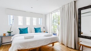 A bedroom with a wooden bed, white bedding, two blue pillows, a bedside table with a plant, and a large window with curtains.