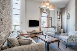 Ein modernes Wohnzimmer mit einer Sofagarnitur, einem Couchtisch und gemusterten Sesseln. Ein Flachbildfernseher ist an der Wand befestigt, und es gibt große Fenster mit Vorhängen.