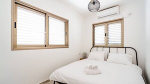 Un dormitorio con cama, dos ventanas con persianas, una mesita de noche y un aire acondicionado de pared.