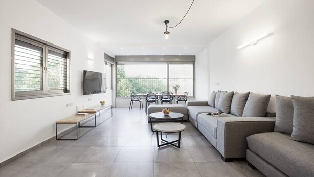 A modern living room with a sofa, a coffee table, and a TV. There is a balcony with a view of trees. - Central 3BR in Rupin by Holiday-rentals SAFE ROOM ({{addressCityy}})