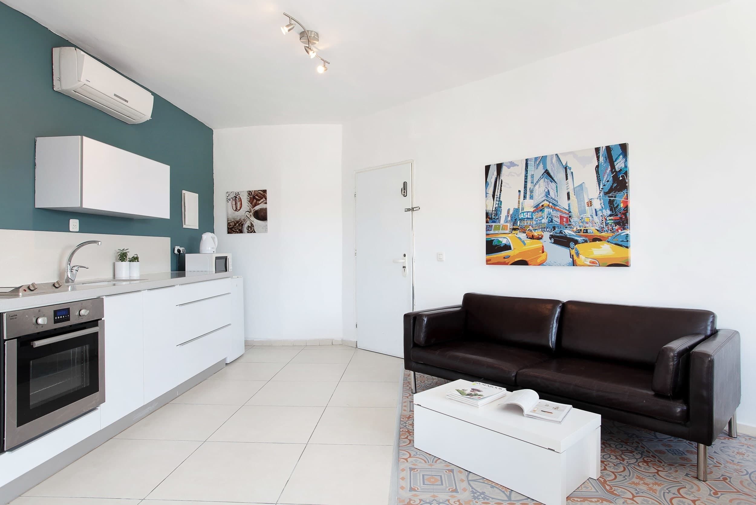 A modern kitchen with white cabinets, a built-in oven, and a microwave. There is a black leather sofa and a coffee table with a book. A painting of a cityscape is on the wall.