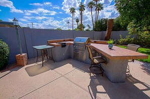 An outdoor kitchen with a grill, a table, and chairs, surrounded by a concrete wall and palm trees.