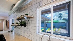 Een keuken met witte tegels, een raam met uitzicht op een zwembad en stoelen, en planken met planten.