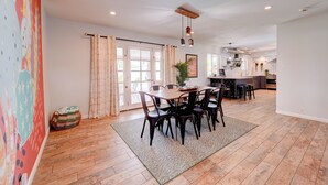 Een ruime eetruimte met een houten tafel en stoelen, een keuken op de achtergrond en een groot raam met gordijnen.
