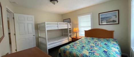 A room with bunk beds, a single bed with a colorful floral bedspread, a small table, a lamp, and a framed picture on the wall.