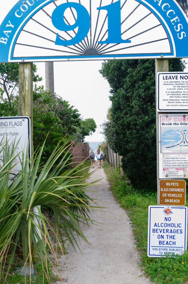 Un sentier menant à l’entrée d’une plage, agrémenté de plusieurs panneaux d’information, dont un panneau indiquant qu’il est interdit de consommer des boissons alcoolisées sur la plage.
