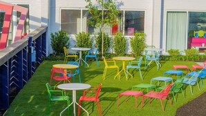 Un área de estar al aire libre con varias mesas y sillas, rodeada por un césped bien cuidado y un edificio con amplias ventanas.