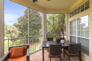 A covered patio with a table and chairs, large windows, and a view of greenery.