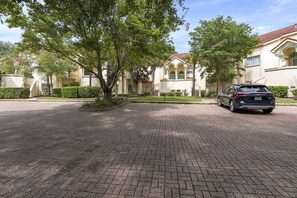A residential street with a paved driveway, a parked car, and houses with arched windows and a tiled roof.