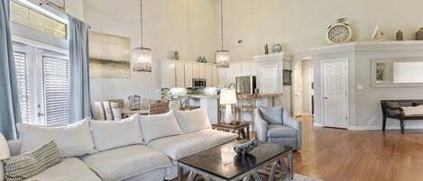 A spacious living room with a white sofa, a coffee table, and a gray armchair. There is a kitchen area with white cabinets and a dining table.