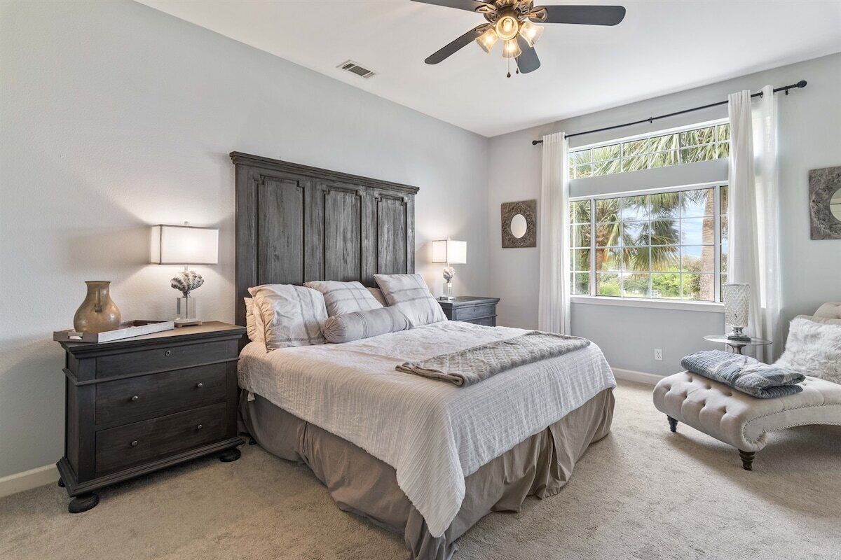 A bedroom with a large bed, a nightstand, a dresser, a chair, a ceiling fan, and a window with curtains.