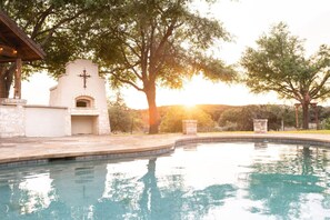 A swimming pool with a stone structure and a cross, surrounded by trees and a sunset.
