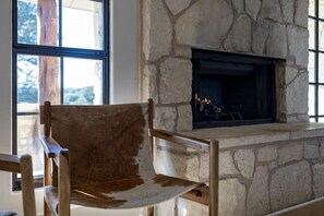 A stone fireplace with a fire, a wooden chair in front, and a window with a view of trees. - Luxury Ranch Retreat Pool, Sleeps 18 (Dripping Springs)