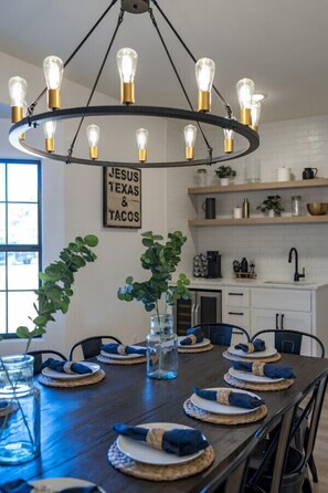 A dining area with a dark wooden table set for six, a modern chandelier, and a wall sign reading 'JESUS TEXAS & TACOS'.