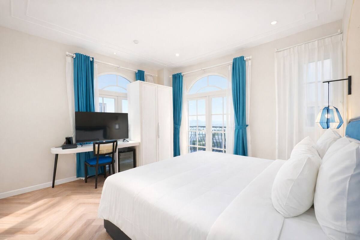 Habitación de hotel con una cama grande, un escritorio con televisor y un balcón con cortinas azules.