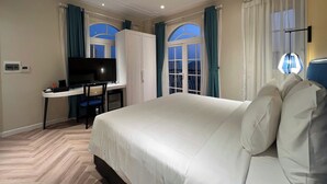 Ein Hotelzimmer mit einem großen Bett, einem Schreibtisch mit Fernseher und zwei fensterbepflanzten Fenstern.