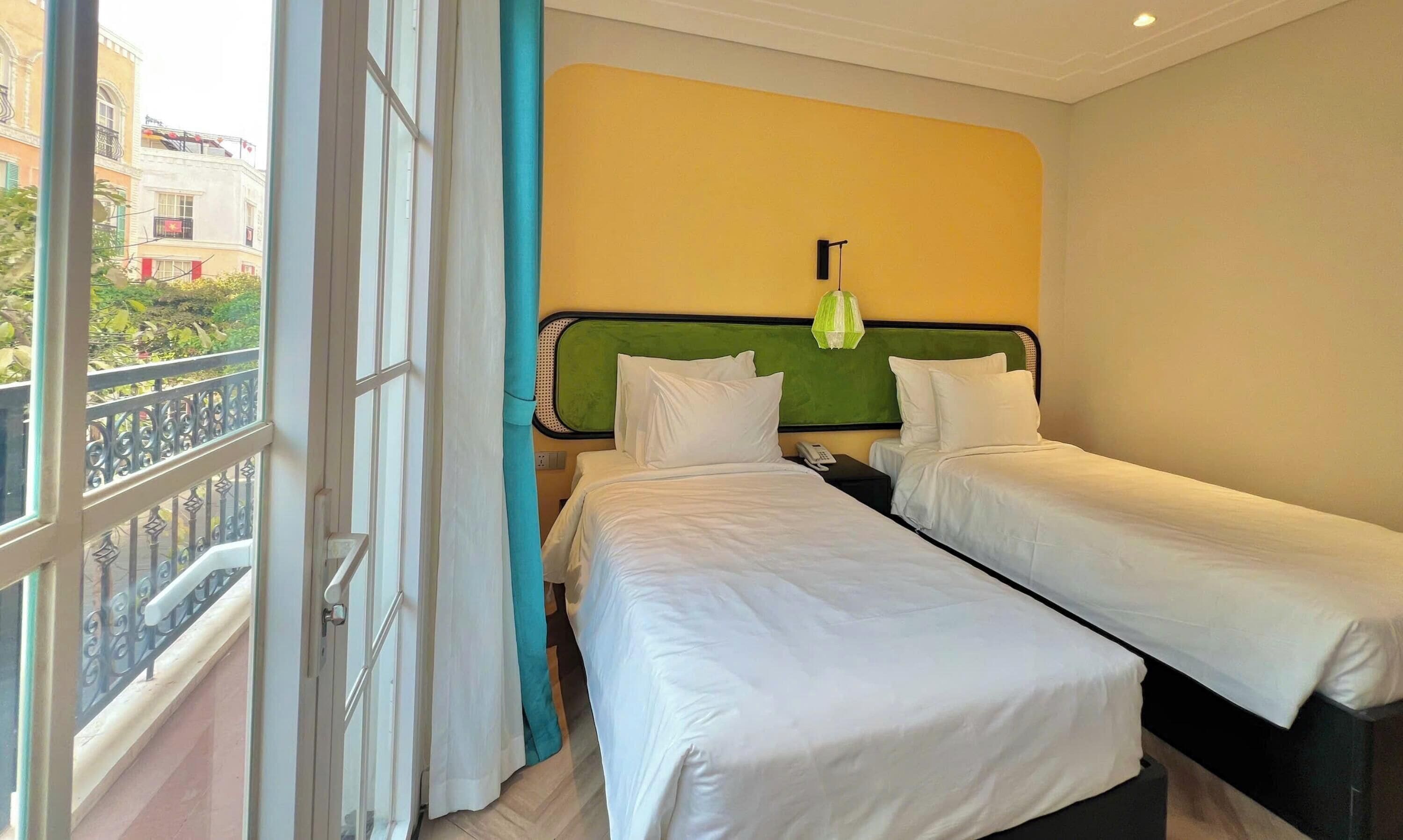 A hotel room with two single beds, a green headboard, and a window with a view of buildings.