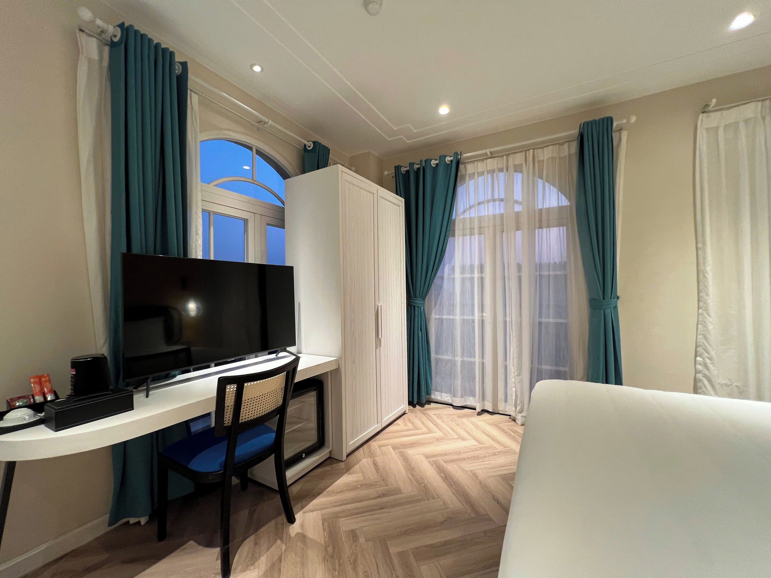 Una habitación moderna con una televisión de pantalla plana, un escritorio y una silla. Se ve una ventana con cortinas y un sofá blanco.
