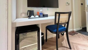 Ein kompakter Raum mit weißem Schreibtisch, einem fest installierten Flachbildfernseher, einem kleinen Kühlschrank und einem Stuhl mit blauer Sitzkissen.