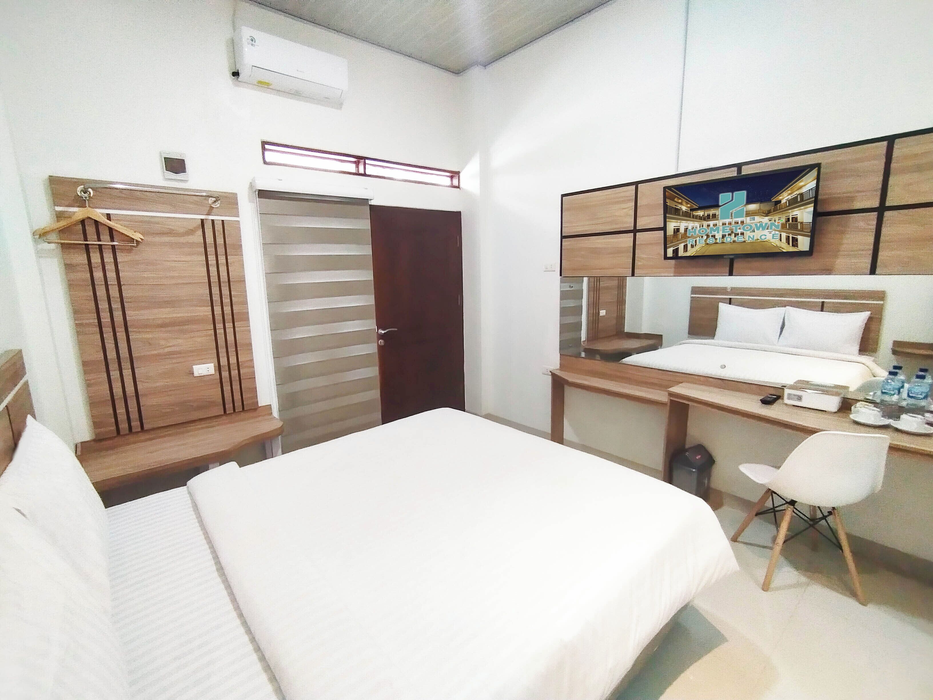 A hotel room with a bed, desk, chair, and a TV mounted on the wall displaying 'HOMETOWN EXPERIENCE'.