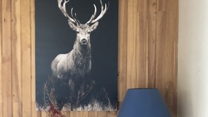 Un mur en bois orné d’un tableau représentant un cerf, une lampe bleue, des bougies, un vase avec des fleurs séchées et une bouteille posée sur une commode en bois.