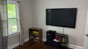 Ett rum med en stor, platt-TV som är monterad på väggen, trägolv, ett fönster med mönstrade gardiner och en liten bokhylla med diverse föremål.