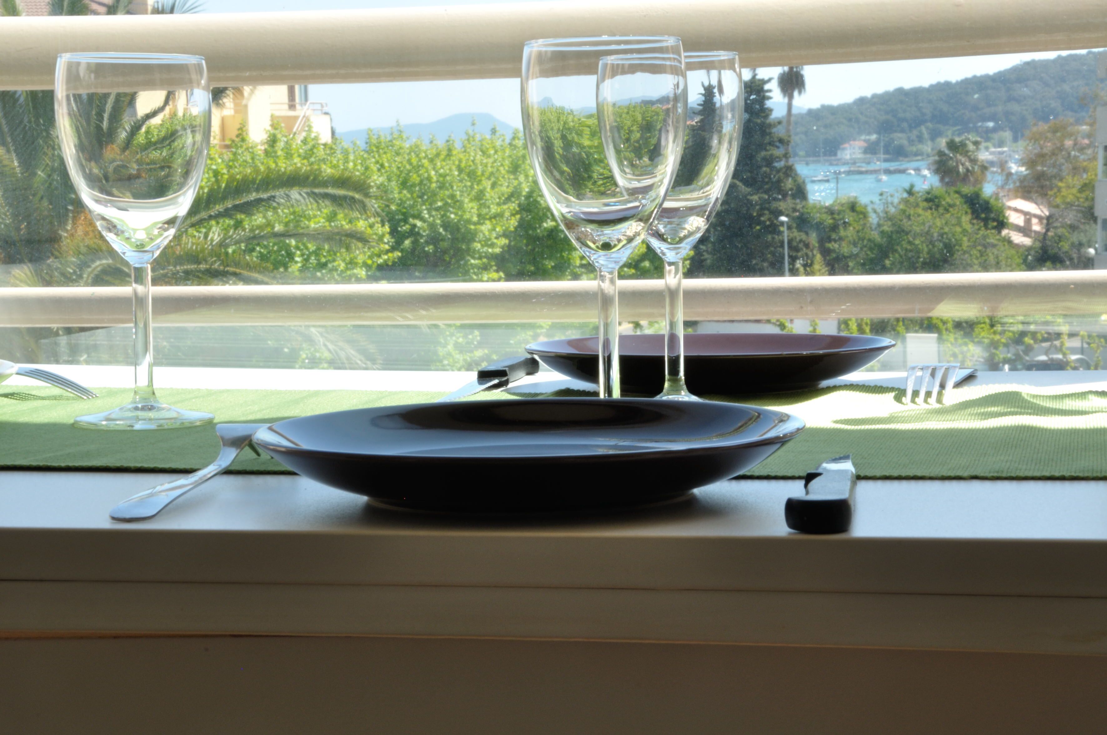 A dining table set with wine glasses, plates, and cutlery, with a view of a coastal landscape through a window.
