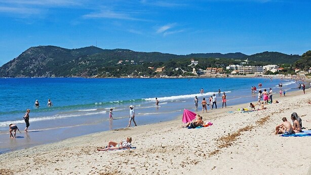 Ein Strand mit badenden Personen, Liegestühlen und Gebäuden im Hintergrund. Ein rosafarbener Sonnenschirm ist ebenfalls zu sehen.