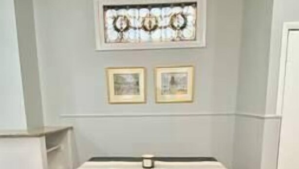 A room with a wooden floor, a table with a white tablecloth, two framed pictures on the wall, and a decorative window above.