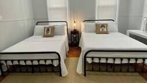 A room with two single beds, each with a white mattress and a wooden headboard. There is a small table between the beds with a lamp on it.
