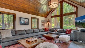 A spacious living room with a large sectional sofa, a coffee table, and a fireplace. There are large windows providing a view of the forest. - Finch Haven in Homewood (Homewood)