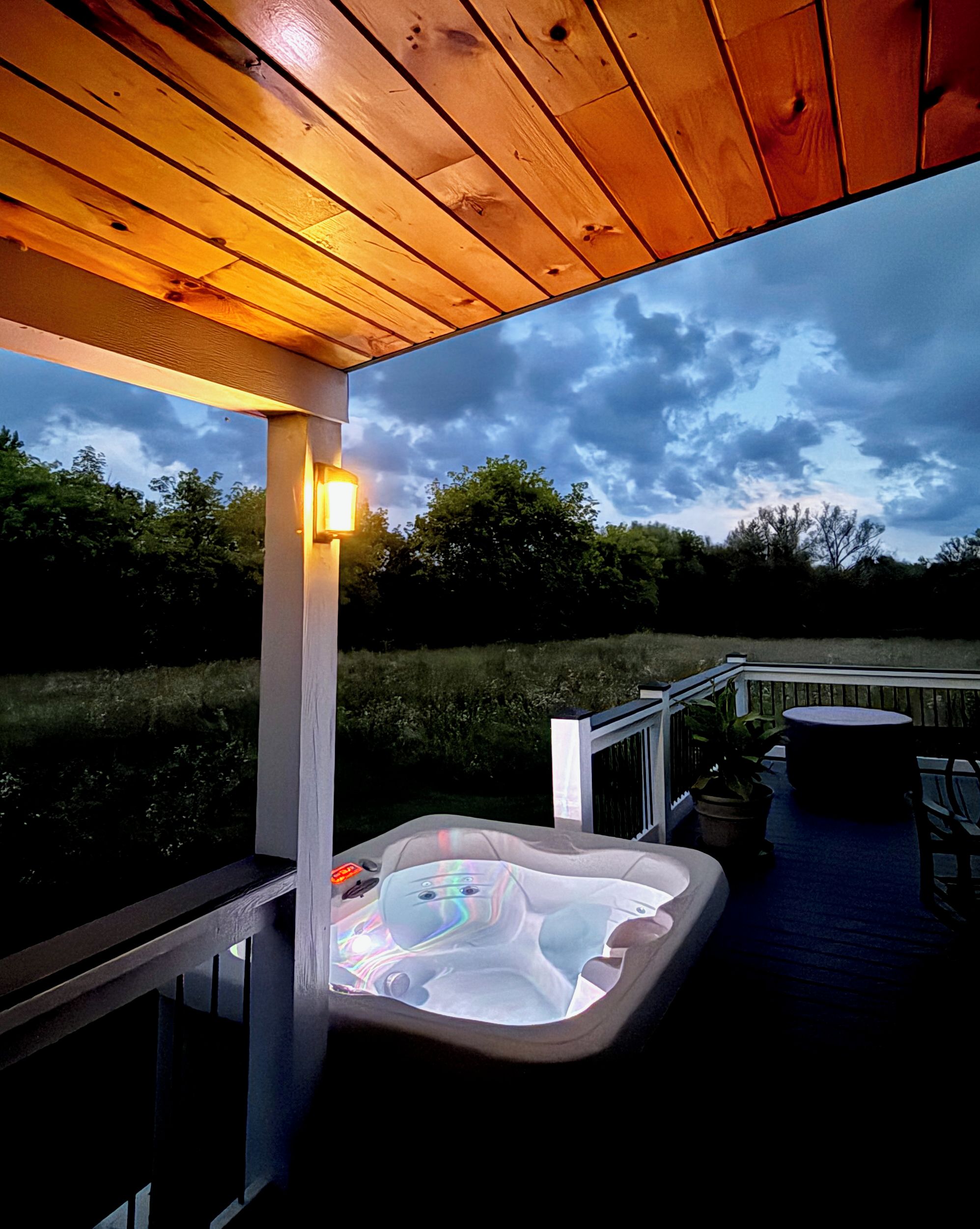 Een hottub op een houten terras met een reling, onder een overdekte ruimte met een lamp.