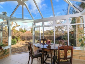 A conservatory with a glass roof, a dining table, chairs, and a view of the backyard. - Oakmead (Moreton-in-Marsh)