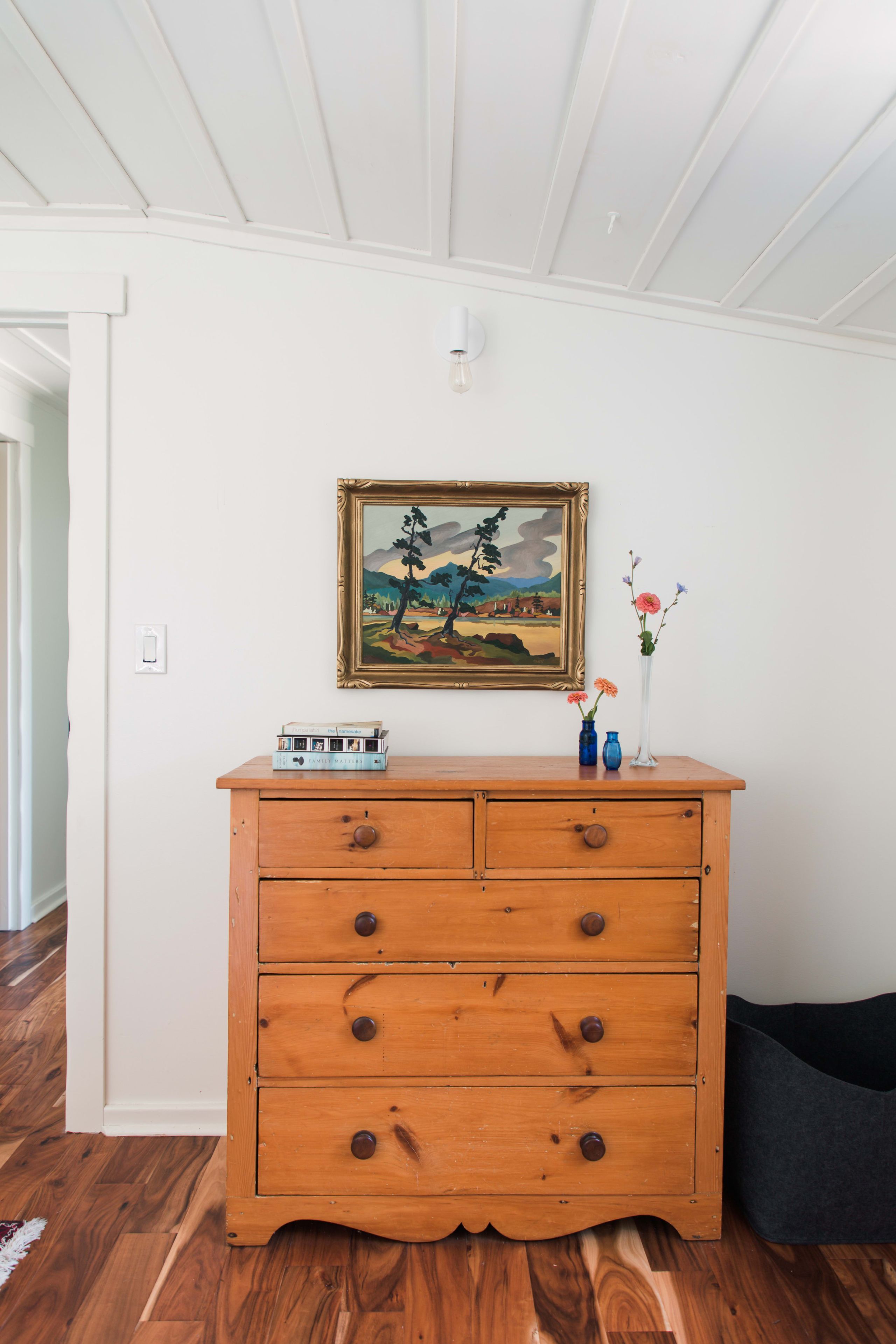 Een houten dressoir met laden, erboven een ingelijst schilderij, en een vaas met bloemen op het dressoir.