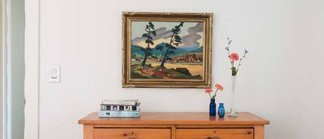 Een houten dressoir met laden, erboven een ingelijst schilderij, en een vaas met bloemen op het dressoir.