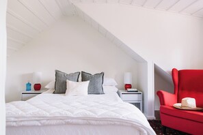 A bedroom with a white bed, grey and white pillows, a red armchair, and a small table with a hat.