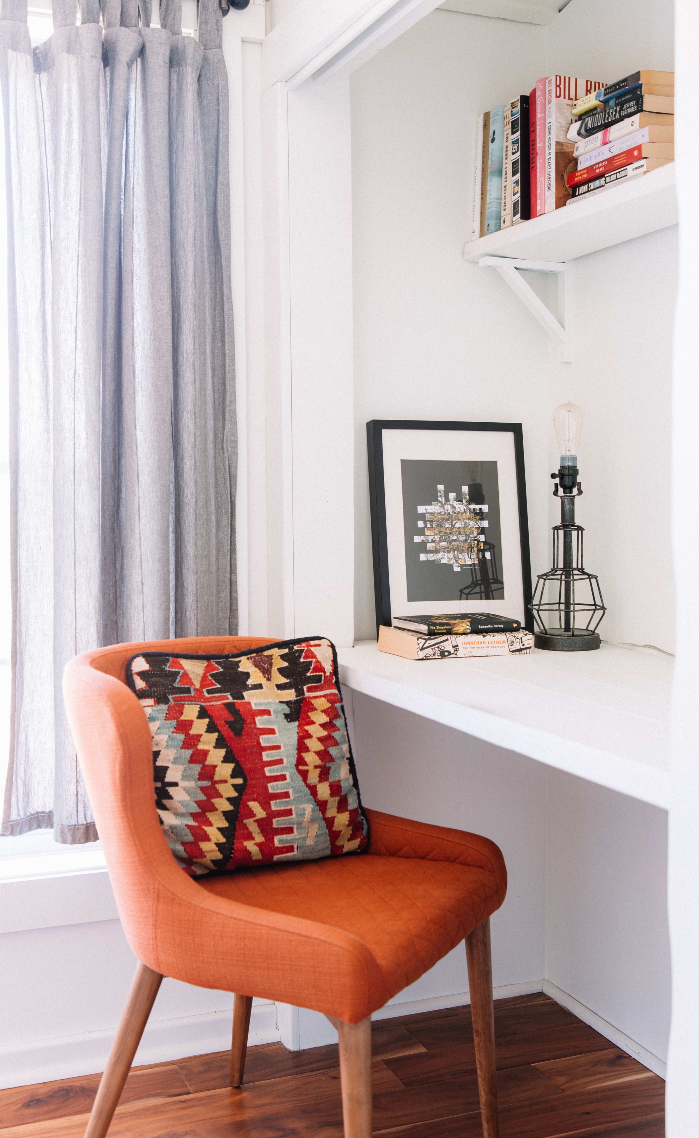 Un coin de pièce avec un plancher en bois, un mur blanc, une fenêtre avec des rideaux gris, une étagère blanche avec des livres et un tableau encadré, et une chaise orange avec un coussin à motifs.