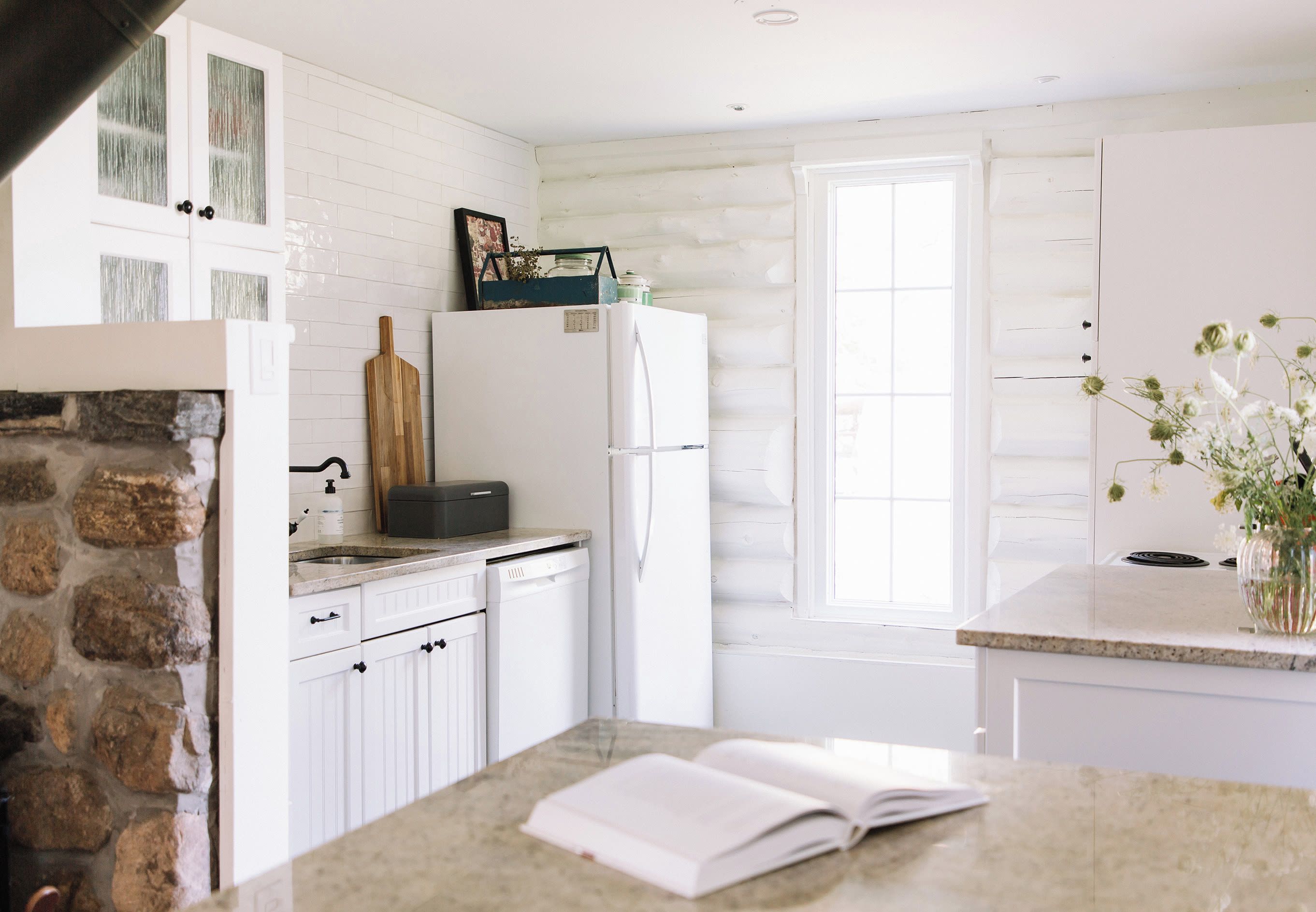 A kitchen with white cabinets, a stone wall, a window, and a countertop with a book.