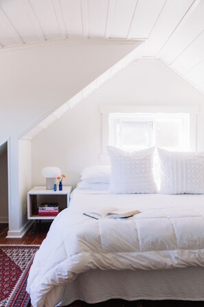 A bedroom with a sloped ceiling, a bed with white bedding, a nightstand with a lamp and books, and a window with natural light.